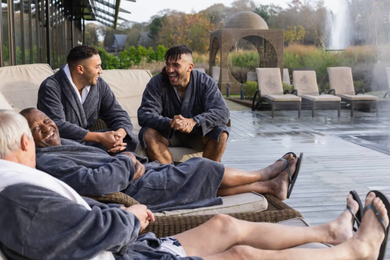 Diverse group of men enjoying a relaxing spa day outdoors, laughing and talking on comfortable chairs in cosy grey robes.